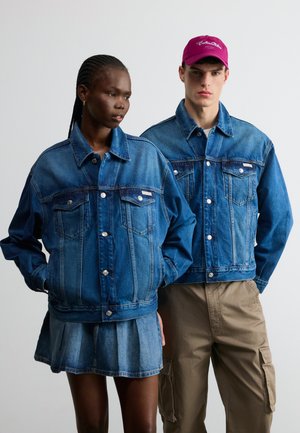 Két modell, akik kék farmerdzsekit viselnek; az egyik farmer szoknyával, a másik bézs terep nadrággal és lila sapkával párosítva.