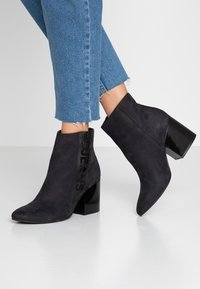 Botas de tobillo de ante negro con un tacón grueso y brillante y un logo en relieve en el costado. El diseño presenta una punta afilada y una textura suave.