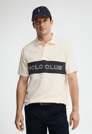 Uomo che indossa una polo color crema con la scritta "POLO CLUB" sul petto, un cappellino blu scuro, pantaloni scuri e una cintura marrone su uno sfondo semplice.