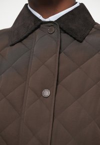 Gros plan d'une veste matelassée marron avec un col en velours côtelé et deux pressions visibles sur un col de chemise blanc.