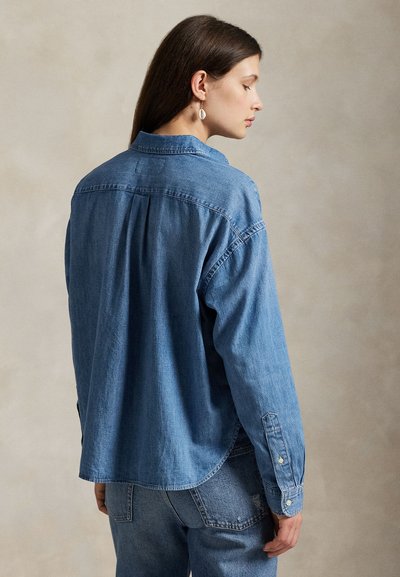 Vrouw met lang bruin haar, die een losse blauwe denim blouse en spijkerbroek draagt, kijkend opzij tegen een effen beige achtergrond.