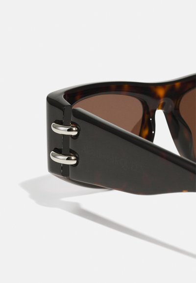 Gafas de sol carey con un marco geométrico grueso. Lentes marrones, acentos metálicos en las patillas y logotipo de marca grabado en el lateral.