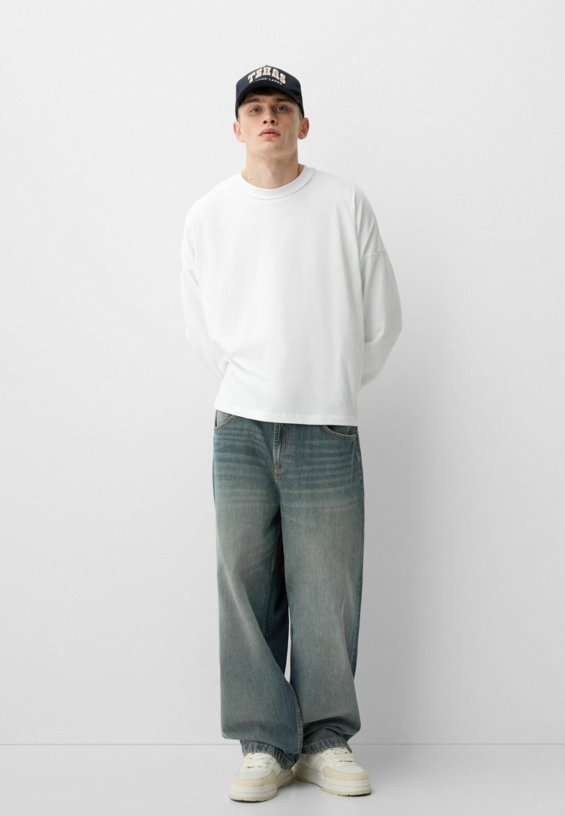 Chemise blanche à manches longues, jean bleu clair déchiré à coupe ample et baskets blanches. Casquette noire avec lettrage blanc, design décontracté.