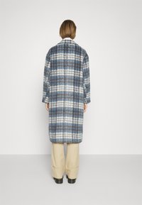 Dlouhý kostkovaný kabát v odstínech modré a bílé, vyrobený z texturované látky. Kabát má volný střih a je kombinován s béžovými kalhotami.