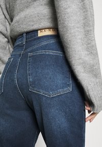 Persona che indossa jeans scuri a vita alta con tasche posteriori e un maglione grigio a maglia, mano appoggiata vicino alla coscia, in un ambiente da studio.