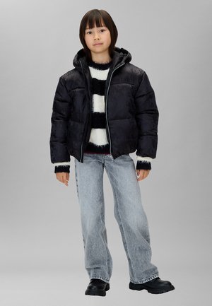 Niño con una chaqueta acolchada negra sobre un suéter a rayas blanco y negro, vaqueros azul claro y botas negras, de pie frente a un fondo gris.