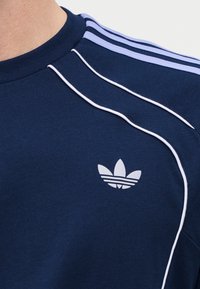 Κοντινό πλάνο σε μπλε σκούρο πουκάμισο Adidas με ανοιχτό μπλε ρίγες στους ώμους, λευκές λεπτομέρειες και το λογότυπο Adidas trefoil στο στήθος.