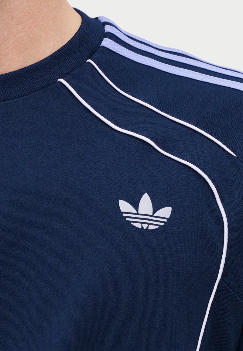 Close-up of navy blue Adidas shirt showing light blue shoulder stripes, white piping, and Adidas trefoil logo on chest.