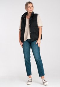 Czarna kamizelka puffy z wysokim kołnierzem, z przednimi kieszeniami, nałożona na beżowy sweter z dzianiny, w połączeniu z niebieskimi dżinsami i wzorzystymi sneakersami.