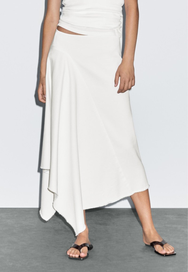 Woman wearing an asymmetrical white midi skirt with raw edges, a white sleeveless top, and black toe-ring flat sandals.