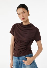 Brun t-shirt med korte ærmer og draperet halsudskæring samt samling på siden, parret med blå denimjeans. Glat stoftekstur.
