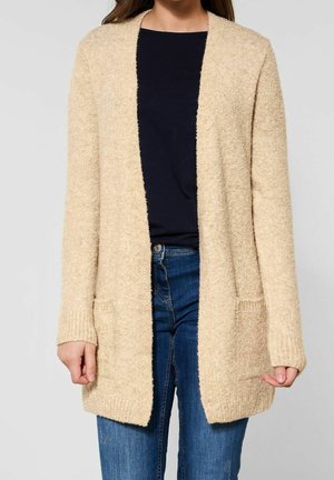 Beige, textured cardigan with long sleeves, an open front, and two front pockets, paired with a dark top and blue jeans.