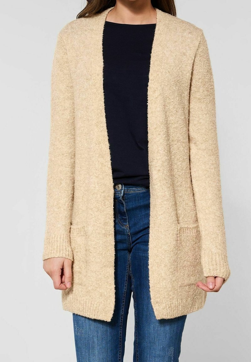 Beżowy, teksturowany kardigan z długimi rękawami, otwartym przodem i dwiema przednimi kieszeniami, połączony z ciemnym topem i niebieskimi dżinsami.