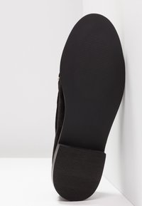 Chaussure en suède noir avec une semelle en caoutchouc texturée, dotée d'un talon bas et d'un bout arrondi et lisse. Accent métallique doré près de l'arrière.
