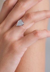 Silberner Herzring mit glitzernder Oberfläche, auf einem schlanken Band, getragen an einer Hand mit glatter Haut.