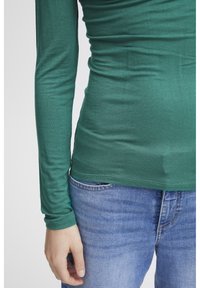 Långärmad grön topp i mjukt tyg, med en figurnära design och rundad nederkant. Kombinerad med blå denimjeans.