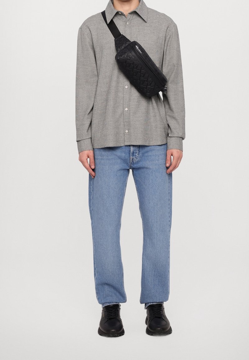 Homme portant une chemise grise à boutons, un jean bleu clair, des baskets noires et un sac bandoulière noir avec un motif embossé sur la poitrine.