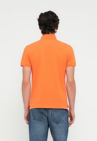 Πορτοκα λί πόλο μπλούζα με γιακά, κοντά μανίκια και πλαϊνές ανοίγματα, συνδυασμένη με τζιν παντελόνι. Λείο, ελαφρύ ύφασμα.