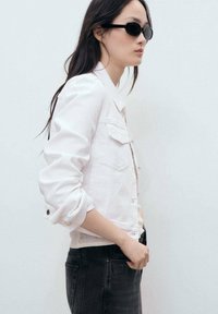 White denim jacket with a structured design, large pockets, and rolled sleeves, paired with black sunglasses and dark pants.