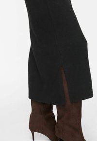 Jupe noire avec fente latérale portée sur des bottes marron en daim à talons hauts, montrée de mi-mollet au genou sur fond blanc.