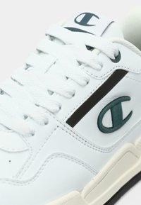 Λευκό δερμάτινο αθλητικό παπούτσι με χοντρές λευκές κορδόνια, σκούρο πράσινο λογότυπο Champion στο πλάι και στη γλώσσα, και κρεμ καουτσούκ σόλα.