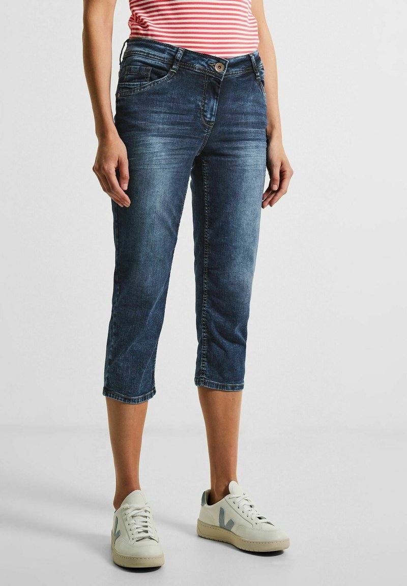 Femme portant un jean court bleu foncé, des baskets blanches et un haut rayé rouge et blanc, debout sur un fond clair uni.