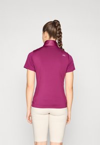 Fitted, kurzärmliges Poloshirt in leuchtendem Magenta mit Kragen, glatter Textur und dezentem Logo-Akzent auf dem oberen Rücken. Kombiniert mit beigen Shorts.