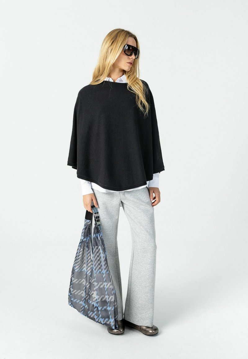 Femme portant un poncho noir, une chemise blanche, un pantalon gris large, tenant un sac fourre-tout à motifs et portant de grandes lunettes de soleil noires, debout sur fond blanc.