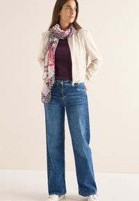 Giacca bomber beige chiaro, top a coste viola, jeans a gamba larga blu e una sciarpa fantasia rosa e grigia. Sneakers bianche completano l'outfit.