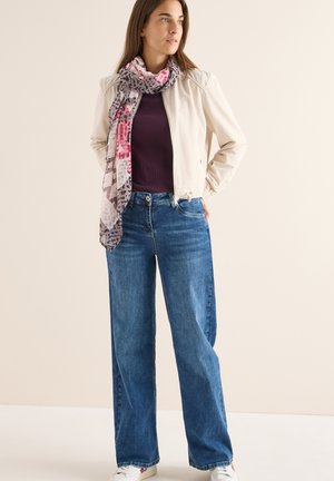 Ljusbeige bomberjacka, lila ribbad topp, vida blå jeans och en mönstrad rosa och grå scarf. Vita sneakers fulländar outfiten.
