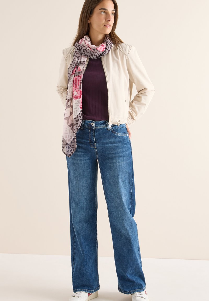 Giacca bomber beige chiaro, top a coste viola, jeans a gamba larga blu e una sciarpa fantasia rosa e grigia. Sneakers bianche completano l'outfit.
