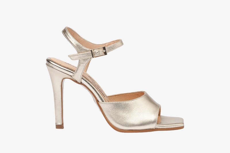 Sandalia de tacón alto metálica dorada con tira en el tobillo, punta abierta, textura suave y plantilla contorneada, que presenta un diseño elegante.