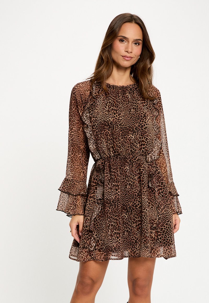 Donna che indossa un abito marrone e nero a stampa leopardata con maniche lunghe arricciate e vita aderente, in piedi contro uno sfondo chiaro e uniforme.