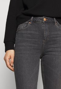 Pantalones vaqueros negros para mujer de corte ajustado, con diseño de cinco bolsillos, botón de cobre y textura sutil. Combinados con un suéter negro.