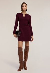 Abito a maglia bordeaux con scollatura a chiave e maniche lunghe, abbinato a stivali in pelle marrone al ginocchio e una piccola borsa nera.