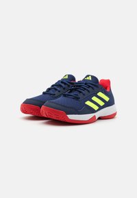 Zapatillas deportivas de color navy con acentos amarillos, parte superior de malla, tacón y suela rojos, patrón de textura en la suela y sistema de cordones tradicional.
