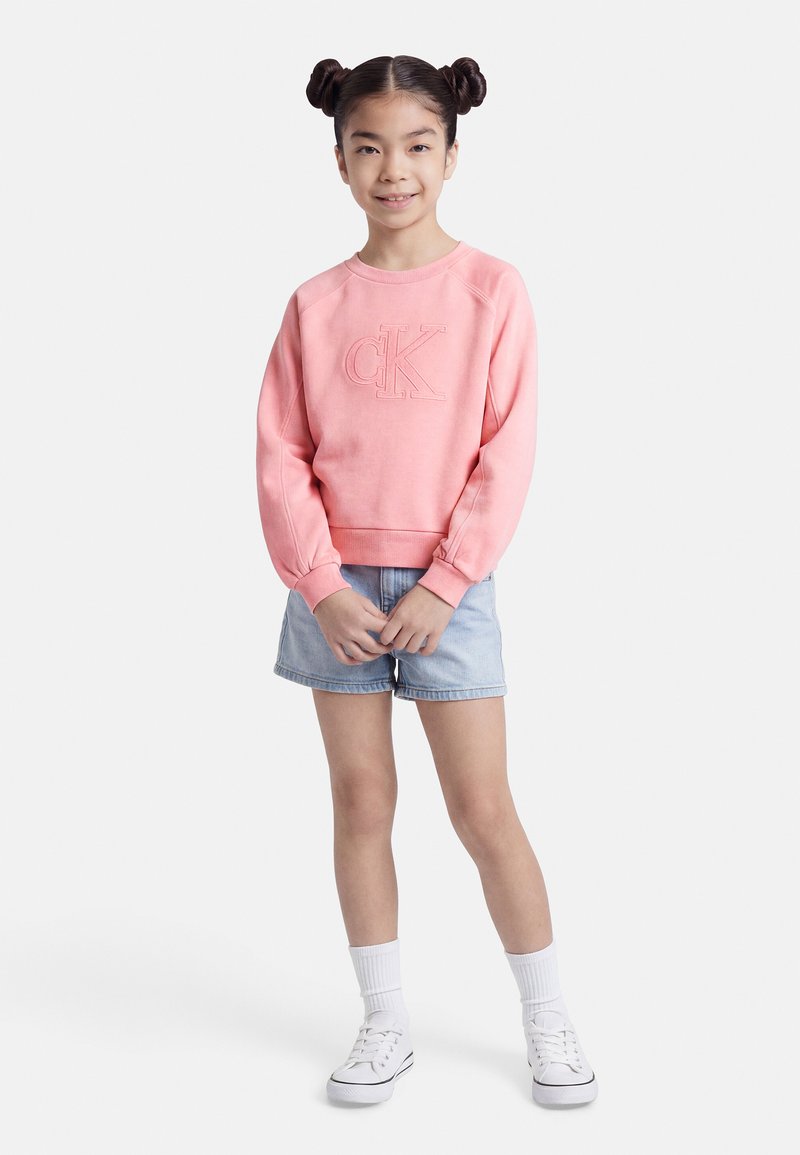 Νεαρή κοπέλα με διπλά σινιόν, φορώντας ροζ φούτερ CK, ανοιχτόχρωμα τζιν σορτς, λευκές κάλτσες και λευκά αθλητικά παπούτσια, στέκεται μπροστά σε λευκό φόντο.