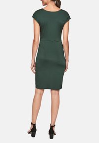 Grön knälång klänning med korta ärmar, figursydd design, slät textur och rundad halsringning. Svarta klackskor kompletterar outfiten.
