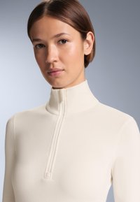 Krémový dlhý rukáv s vysokým zipsom, vyrobený z hladkej tkaniny, s minimálnym šitím a priliehavým strihom.