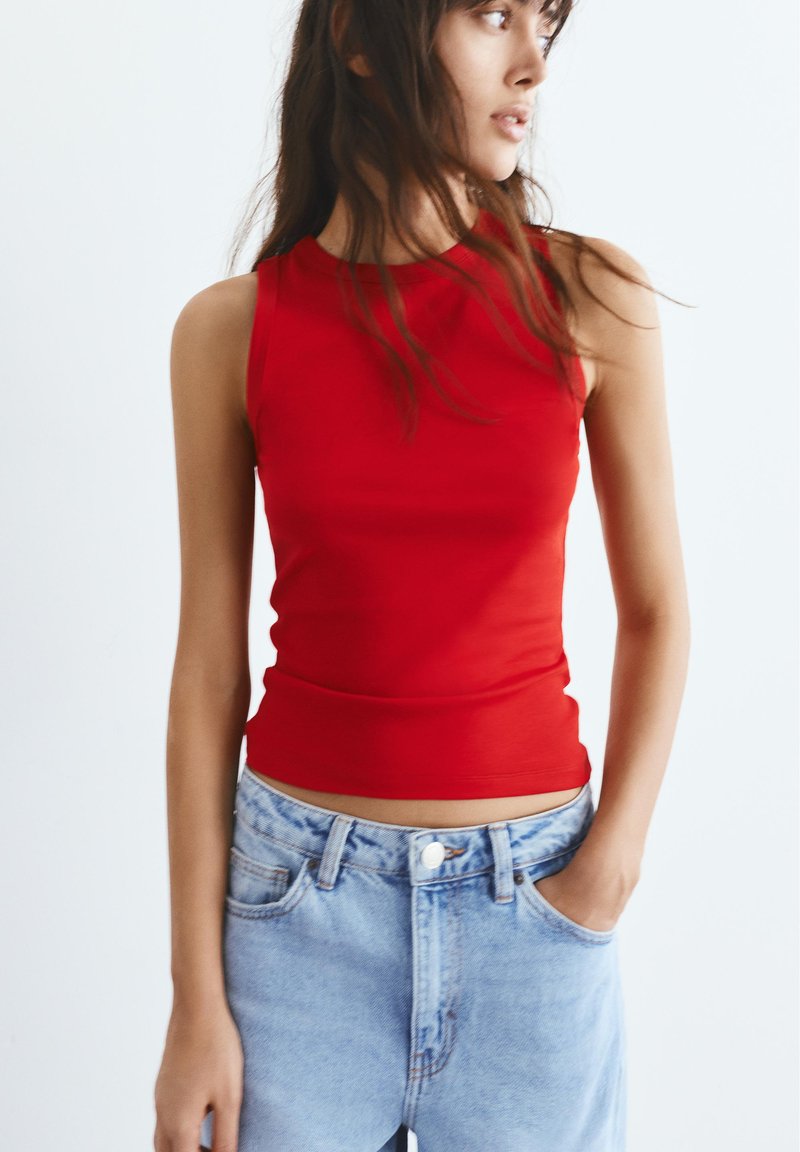 Femme aux cheveux longs portant un haut rouge sans manches et un jean en denim bleu clair, regardant sur le côté avec une main dans sa poche.