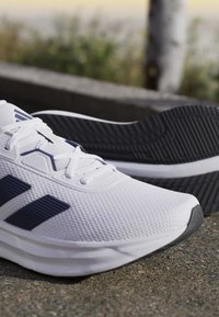 White athletic shoes with black stripes and textured mesh, laced up, placed outdoors on rough pavement with a blurred natural background.