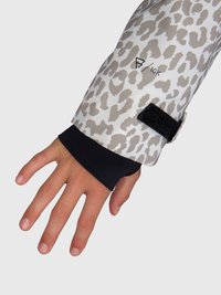 Ruka vystupujúca z rukáva kabátu s leopardím motívom, s čiernym vnútorným manžetou a detailom zapínania na suchý zips na jednofarebnom pozadí.