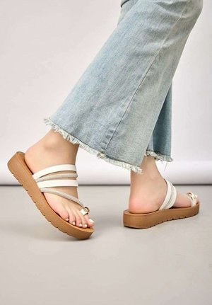 Pieds portant un jean bleu clair à ourlet effiloché et des sandales compensées beiges avec plusieurs fines lanières et un détail en perles dorées.