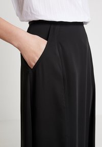 Primo piano di una persona con la mano in tasca, che indossa una gonna nera a vita alta e una camicia bianca su uno sfondo semplice.