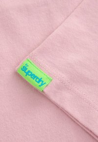 Šviesiai rožinė medvilninė marškinėliai su žaliu etiketu, ant kurio mėlynai išsiuvinėtas užrašas "superdry". Savybės: trumpi rankovės ir minkšta tekstūra.