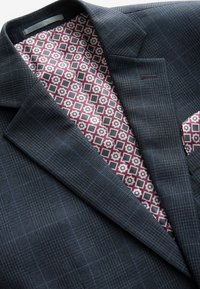 Navy kariertes Blazer aus strukturiertem Stoff. Das Futter zeigt ein geometrisches Muster in Burgunderrot und Grau. Mit einem Akzent in Form eines Taschentuchs.