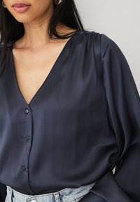 Marineblå silkebluse med V-hals, knaplukning, og bløde draperede ærmer. Stoffet har en glat tekstur og en subtil glans. Bæres med en denim-talje.