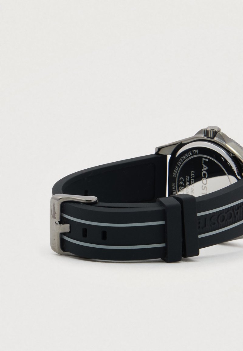 Zwarte rubberen horlogeband met zilveren gesp en gegraveerd Lacoste-logo, bevestigd aan een horlogekast van roestvrij staal.