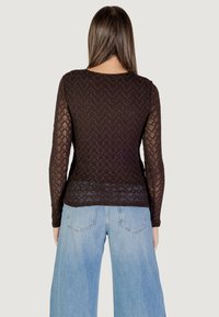 Top lungo in pizzo nero con maniche lunghe, caratterizzato da un motivo a zigzag, indossato sopra jeans a gamba larga azzurri. Vista da dietro.