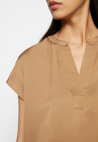 Kurzärmlige Bluse mit V-Ausschnitt aus einem glatten, beige-farbenen Stoff. Lässt sich durch einen subtilen Glanz und ein minimalistisches Design mit einer kleinen Knopfleiste und Stickdetails auszeichnen.